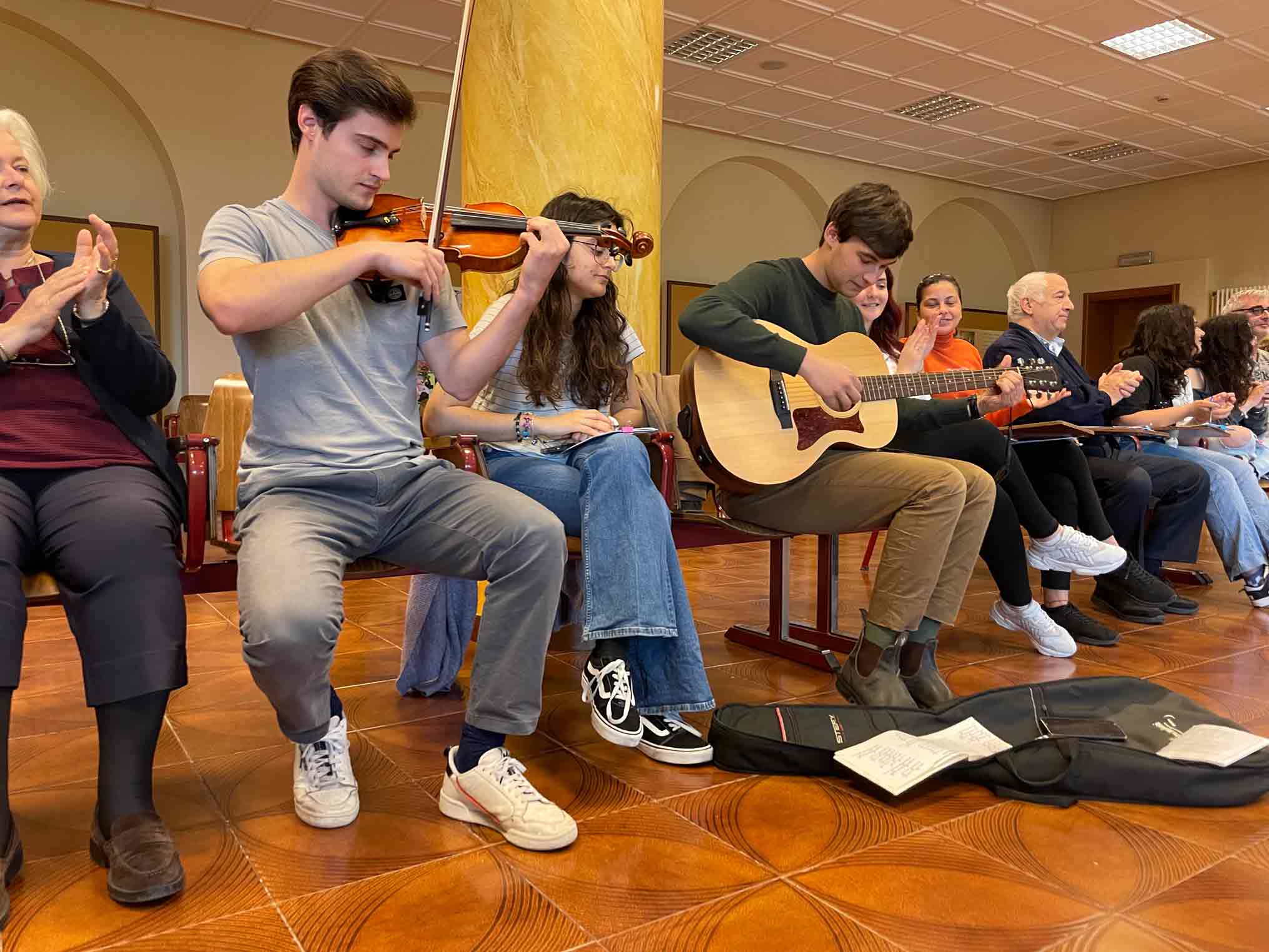 Due ragazzi suonano il violino e la chitarra accanto ad altre persone sedute