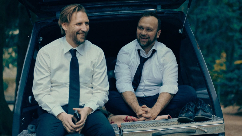 Due uomini con camicia e cravatta ridono seduti nel portabagagli di un auto